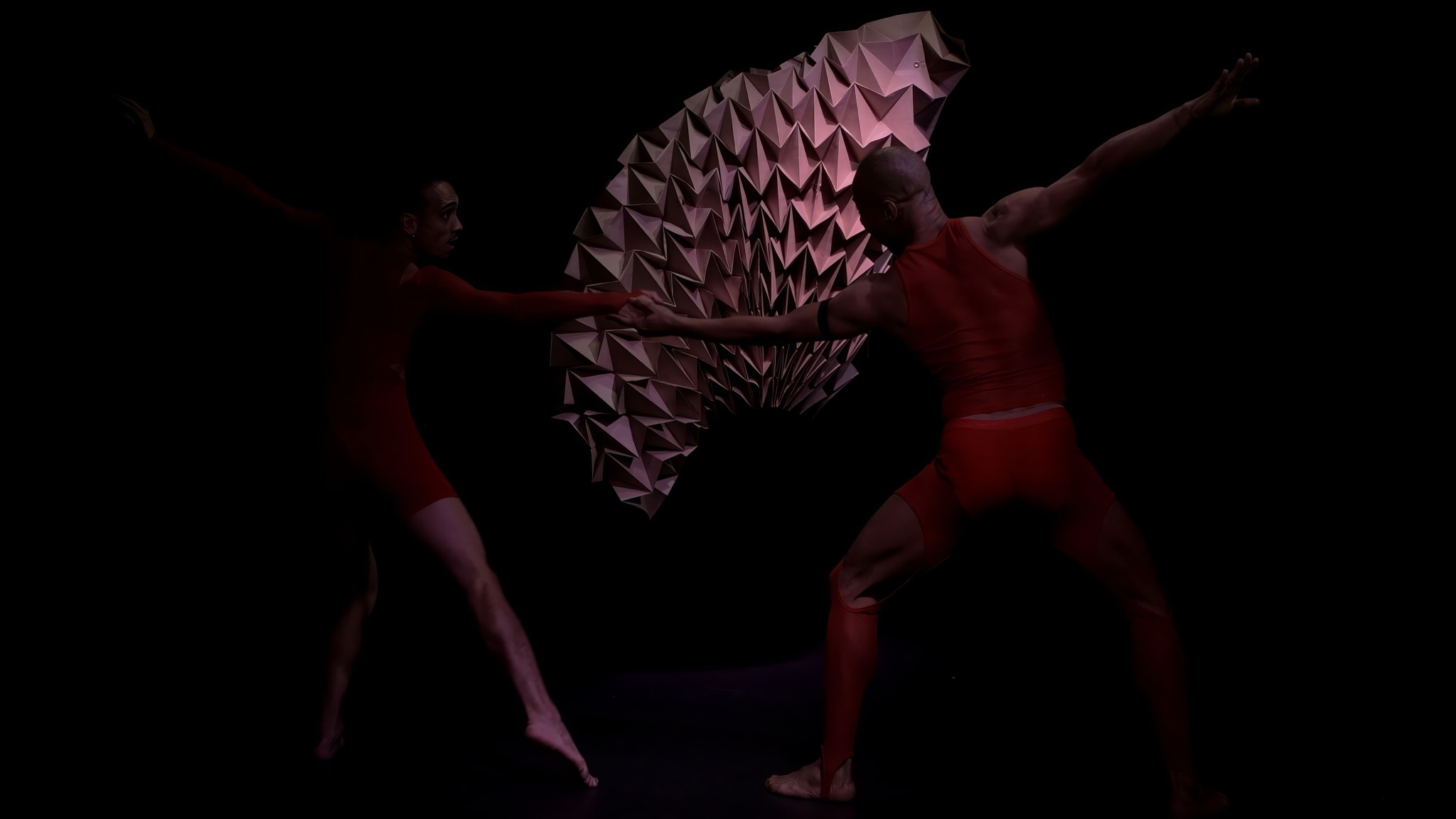 The two dancers in a wide duet in front of the paper sculpture.
